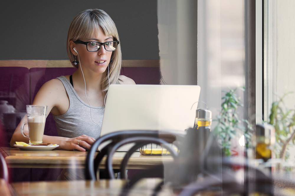Bring your own device. Eine Frau sitzt in einem Café, sie trägt eine Brille, Kopfhörer und arbeitet an ihrem Laptop.