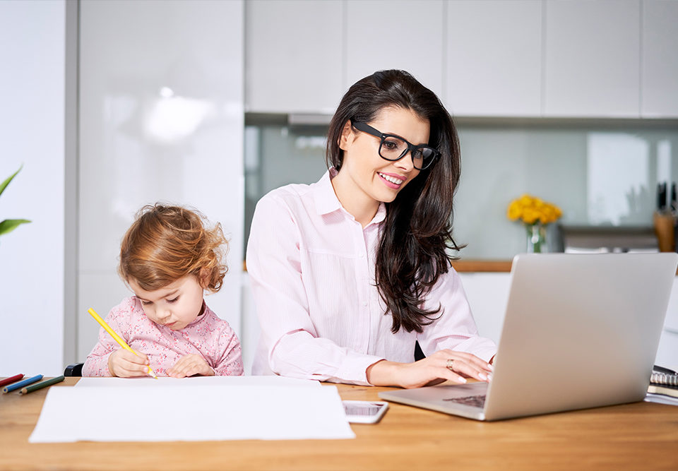 Homeoffice mit Kind. Eine Frau arbeitet, während ein Kind malt.