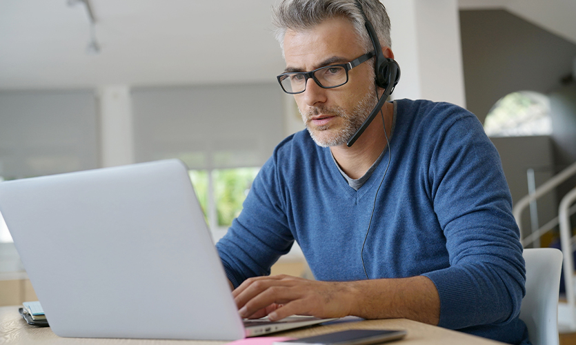 Ein Mann mit Headset und Brille sitzt vor seinem Laptop und arbeitet von Zuhause.